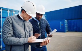 Two engineers standing by freight containers on a large commercial dock discussing shipping logistics together